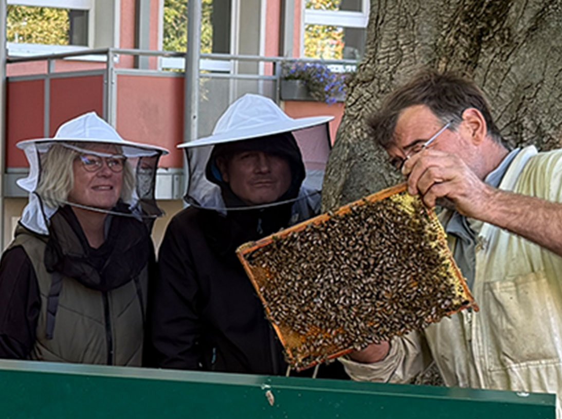S. Fragge und C. Hecht schauen mit Imkerhut dme Imker zu