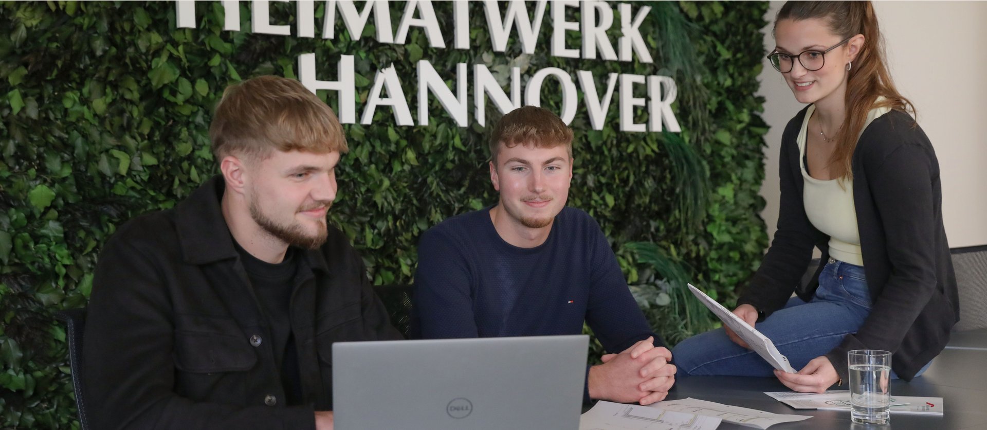 Zwei junge Männer und eine junge Frau sitzen vor Heimatwerk Logo an bewachsener Wand